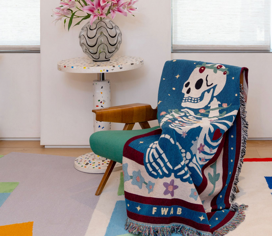 A colorful throw blanket featuring a graphic design with a skeleton, stars, and geometric shapes, draped over a chair in a room with a decorative vase on a table and a colorful floor.