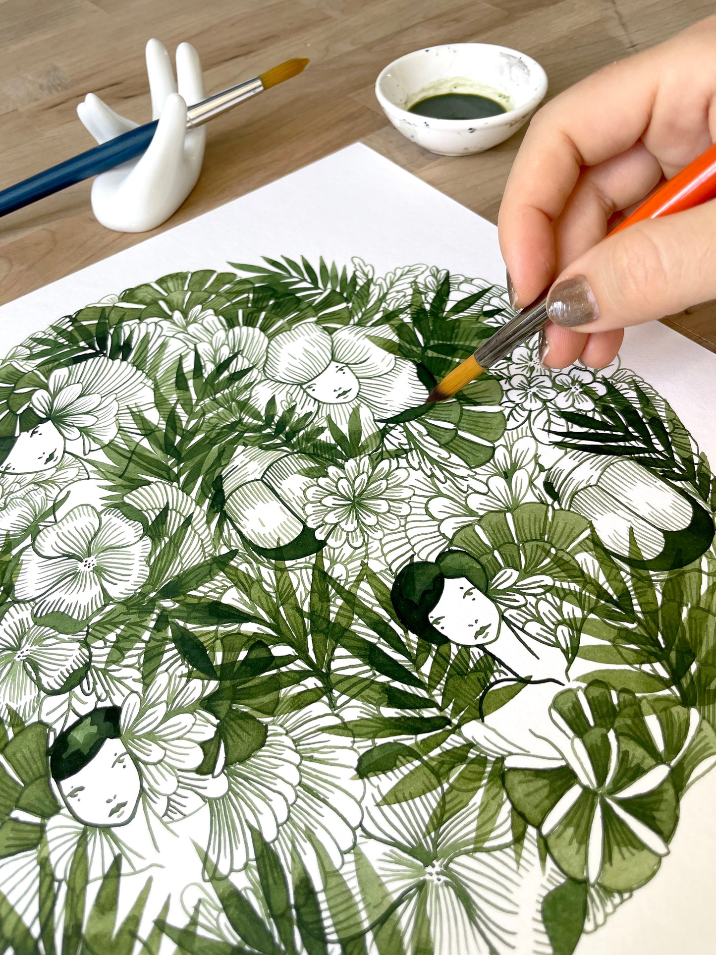 Person painting a detailed illustration of a woman's face surrounded by leaves and flowers on a piece of paper.