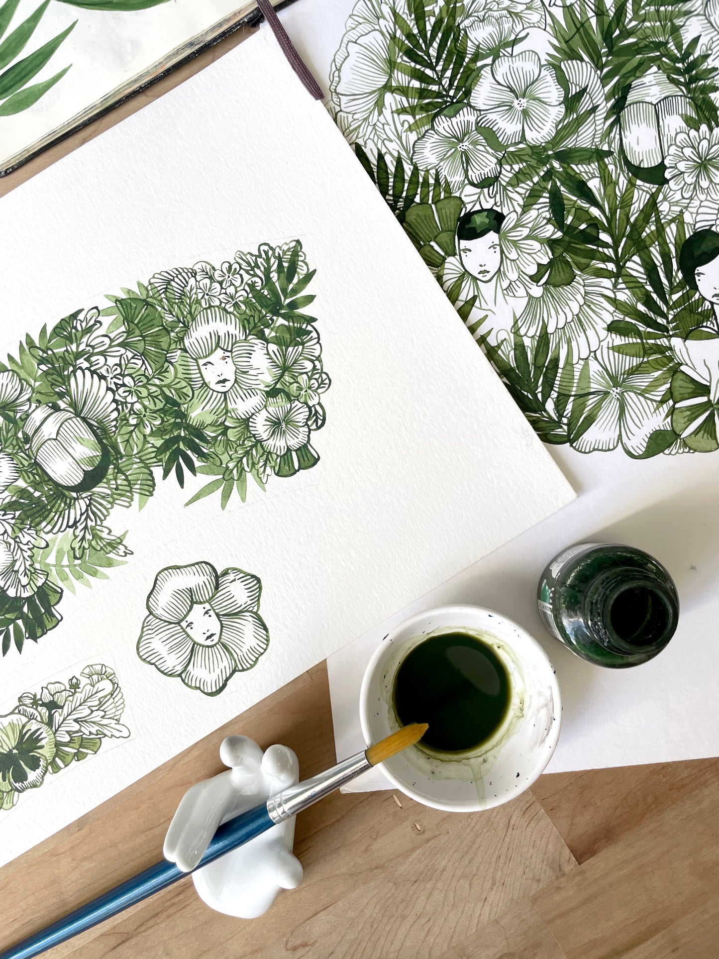 Artistic illustrations of floral and botanical designs on paper with a cup of green liquid and brush.