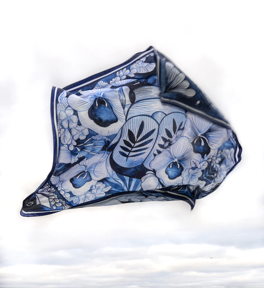 A silk scarf with a floral and animal pattern in shades of blue and white, displayed against a sky-like background.