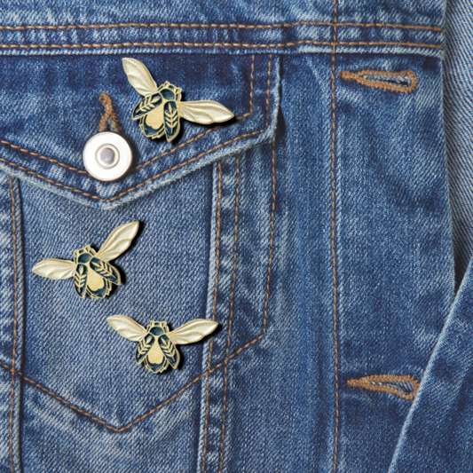 A series of metallic pins with floral and bee designs attached to a denim fabric.