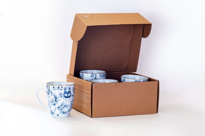 A set of four blue and white illustrated mugs with floral and animal designs, displayed in an open brown cardboard box.