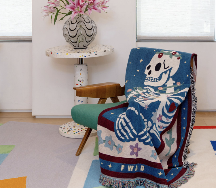 A colorful throw blanket featuring a graphic design with a skeleton, stars, and geometric shapes, draped over a chair in a room with a decorative vase on a table and a colorful floor.