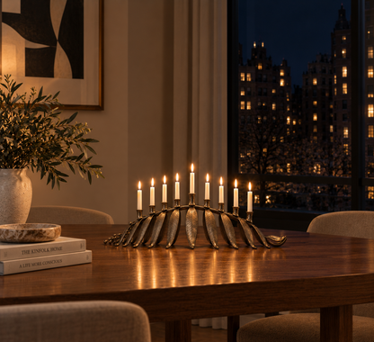 A metallic candelabra with multiple lit candles sits on a wooden dining table in a warmly lit interior, with a window showing a city skyline at night in the background.