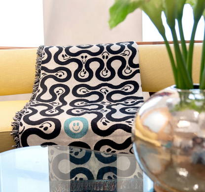 A throw blanket with a geometric pattern featuring black and white colors and blue accents, draped over the arm of a sofa, with a glass vase with a plant in the foreground.