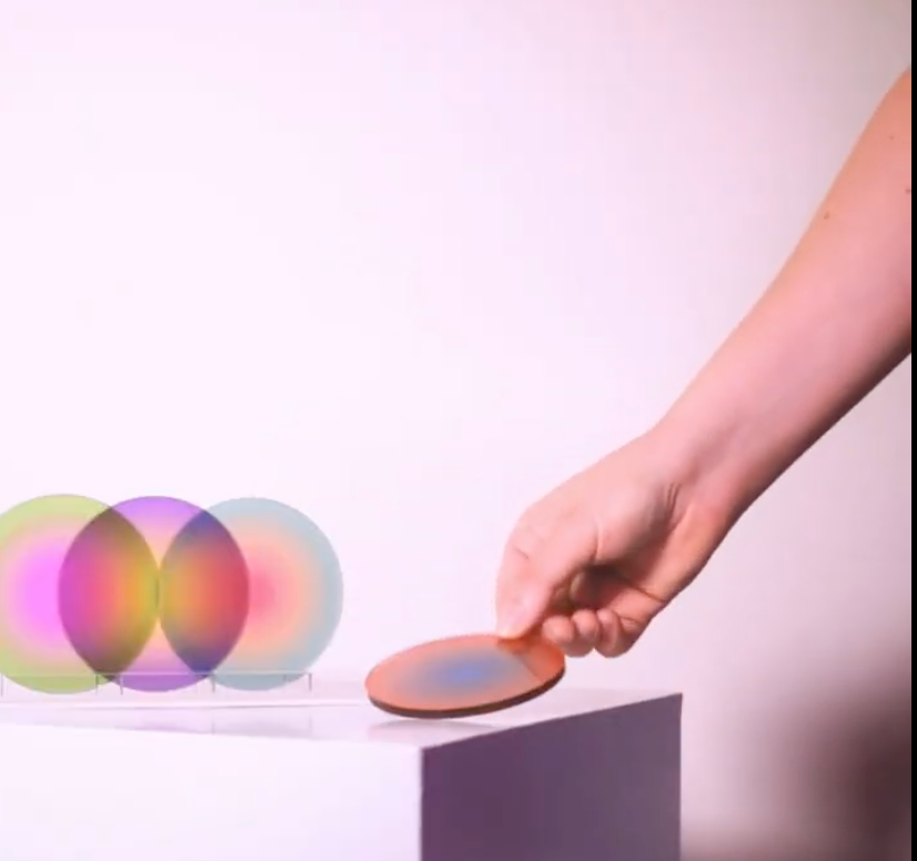 Hand placing a colorful gradient acrylic coaster on a white pedestal beside a clear display stand holding three overlapping gradient coasters designed by graphic artist Yoni Alter.