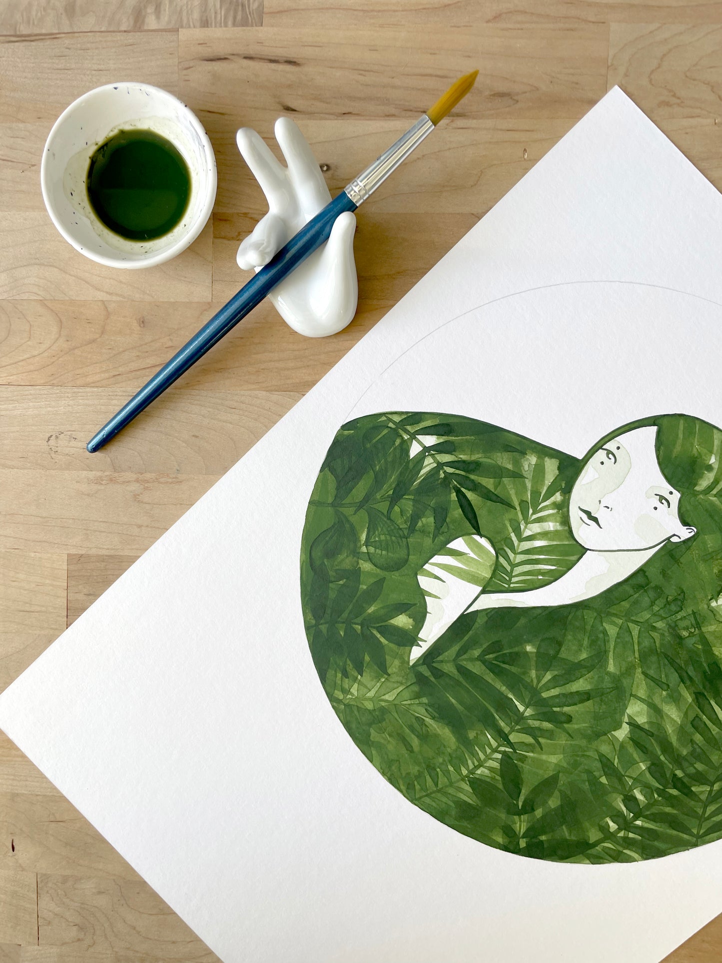 Artistic depiction of a person embraced by leaves on a white sheet of paper, with a cup of green liquid and brush on a wooden surface.