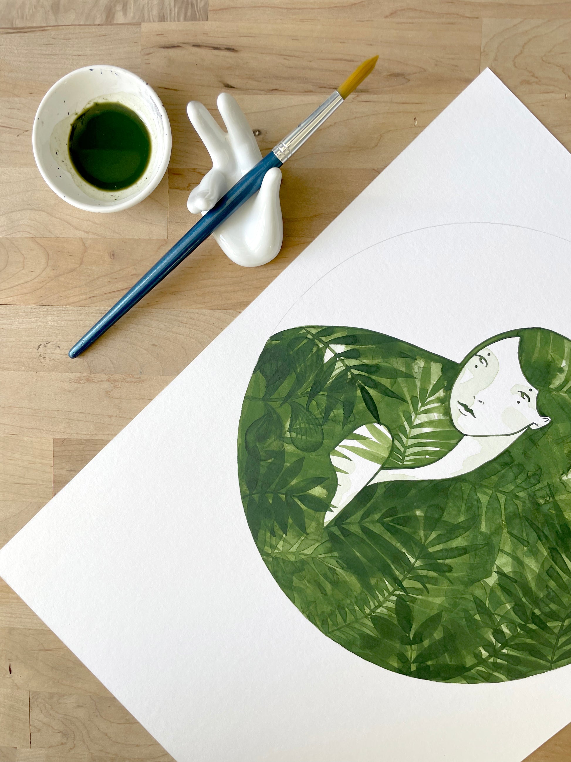 Artistic depiction of a person embraced by leaves on a white sheet of paper, with a cup of green liquid and brush on a wooden surface.