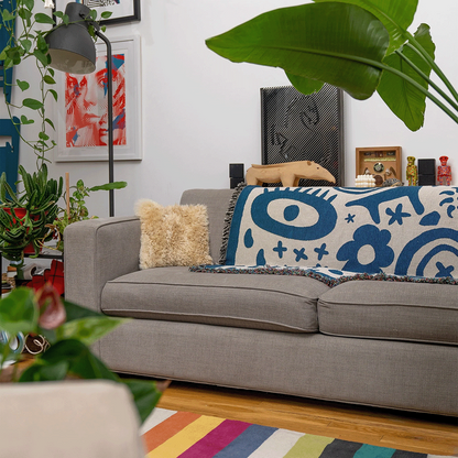 A colorful throw blanket with blue doodles design draped over a gray sofa, in a room with decorative plants, framed art, and a floor lamp.