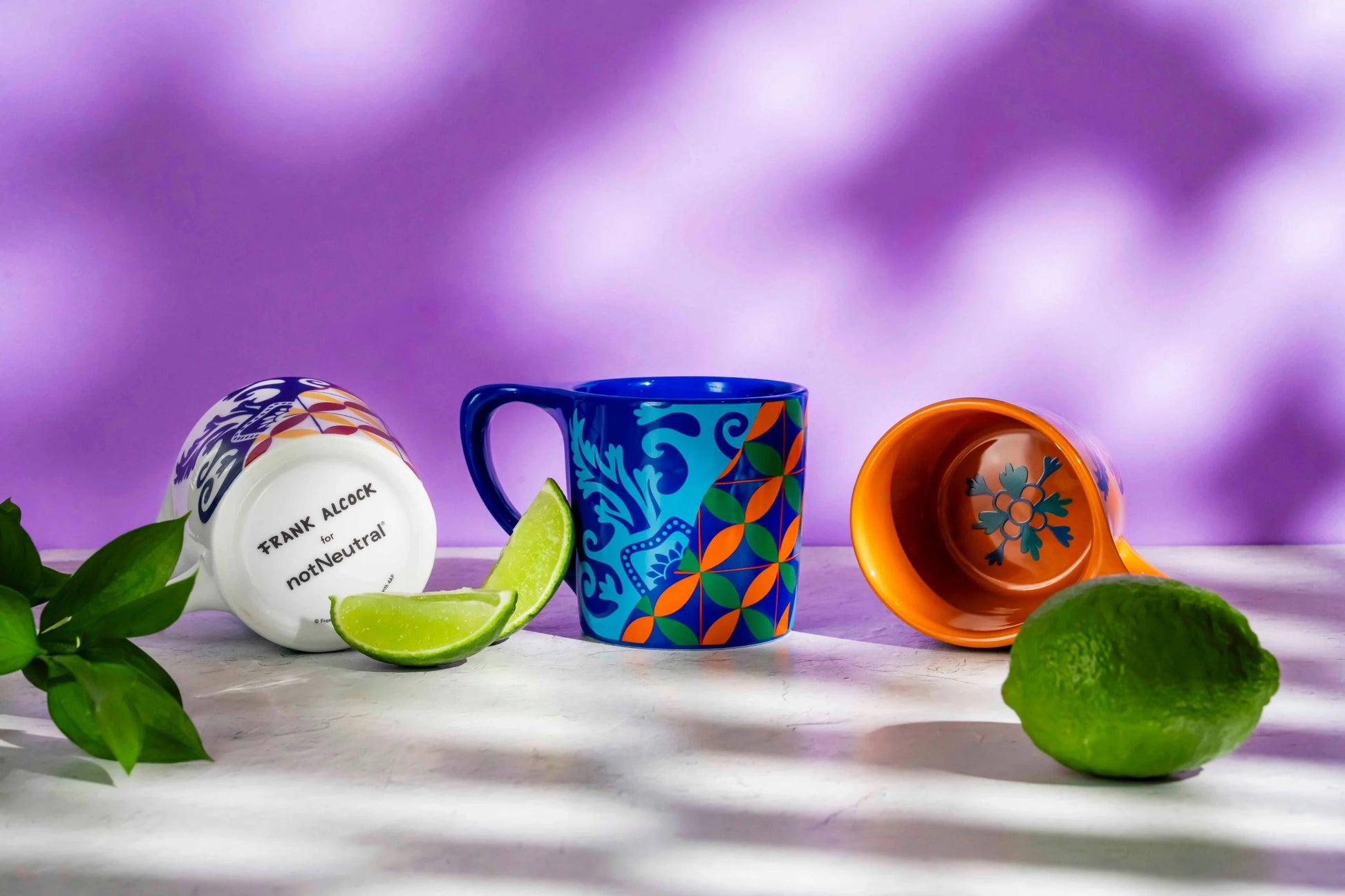 Colorful mugs with decorative patterns on a marble surface with limes and leaves, against a purple gradient background.