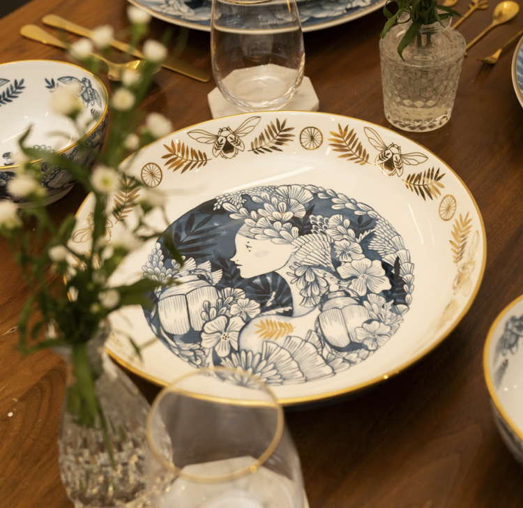 A ceramic serving bowl with a detailed, whimsical illustration of plants and mythical creatures in a monochrome color palette, with gold accents along the rim.