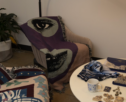 A throw blanket with a bold graphic design featuring abstract faces in purple and black colors, displayed on a chair next to a table with various decorative items.